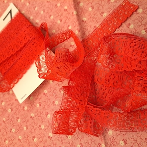 May include: Red lace trim with a delicate pattern. The lace is shown on a pink floral fabric background.