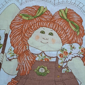 Puede incluir: Un panel de tela vintage con una ilustración de muñeca Cabbage Patch Kids. La muñeca tiene cabello rojo atado con lazos verdes, ojos verdes y viste un vestido con peto marrón con cuello y mangas estampadas florales. El panel incluye instrucciones y la palabra "Cabbage".