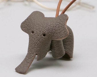 Peut inclure: Un porte-lunettes en forme d'éléphant en cuir gris avec une boucle en cuir marron. L'éléphant a deux petits trous pour les yeux et une surface texturée. Des lunettes sont visibles en arrière-plan.