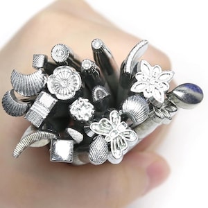 Pode incluir: Uma coleção de ferramentas de estampagem de metal em tom prateado com várias cabeças em relevo, incluindo formas de borboleta, padrões florais, quadrados geométricos, círculos, meias-luas e designs texturizados em forma de gota.