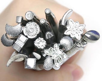 May include: A collection of silver-toned metal stamping tools featuring various embossed heads, including butterfly shapes, floral patterns, geometric squares, circles, crescents, and textured teardrop designs, all bundled together in a cluster.