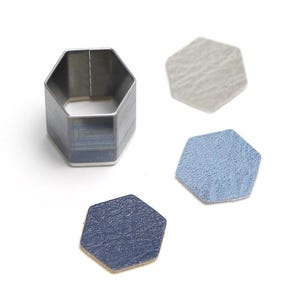 Op de afbeelding: Een metalen stansgereedschap in een zeshoekige vorm, met drie bijpassende zeshoekige stukken van getextureerd materiaal in lichtblauw, donkerblauw en grijs. Het gereedschap is van zilverkleurig metaal.