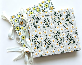May include: Two square fabric-covered albums with white ribbon ties. The front album features embroidered white daisies with yellow centers and green stems on a beige background. The back album displays embroidered yellow sunflowers with green leaves.