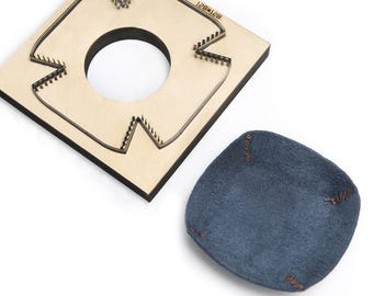 Op de afbeelding: Een vierkant houten stansgereedschap met een centrale ronde uitsparing en een metalen mes, vergezeld van een klein, blauw, vierkant stuk leer met gestikte hoeken, voorzien van de tekst 128*128 gegraveerd op het houten oppervlak.
