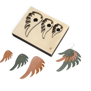 May include: A rectangular metal die cutter featuring three wing-shaped cutouts of varying sizes, marked with the number 70. Beside it are several textured leather wing-shaped cutouts in copper and dark green, plus one assembled earring.