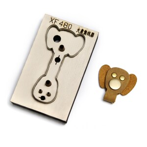 May include: A rectangular metal cutting die labeled XF 480 with an elephant-shaped outline and internal circular punches, alongside a finished tan leather elephant-shaped cord organizer featuring two small brass rivets and one large central brass snap.