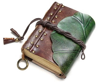 May include: A brown leather journal featuring a green leaf-shaped cover overlay, secured with a braided leather cord. The spine is accented with small brass-coloured metal studs, and a circular metal ring is attached to the bottom corner.