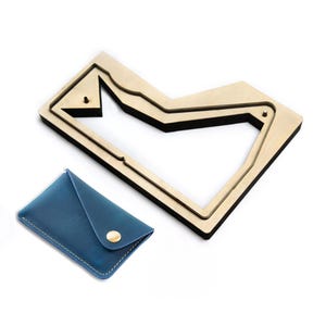 Könnte beinhalten: Ein blauer Kartenhalter aus Leder mit goldfarbenem Druckknopfverschluss und weißen Nähten, begleitet von einer Stanzform aus Holz mit schwarzer Schaumstoffkontur, die das Muster des Kartenhalters bildet, inklusive zwei kleiner Metallstifte.