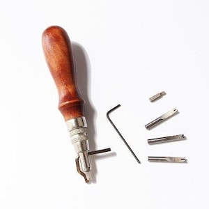 May include: A leathercraft tool set featuring a wooden handle, metal components, and interchangeable blades. Includes an Allen wrench and several blade attachments for various leatherworking tasks. The handle is approximately 5 inches long.