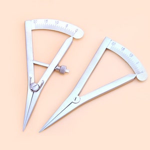 Puede incluir: Dos calibradores de metal plateado con escalas de medición curvas. Cada calibrador tiene una punta y un tornillo ajustable. Las escalas están marcadas con números del 0 al 20. Los calibradores están diseñados para mediciones precisas.