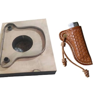 Könnte beinhalten: Ein Holzstempel für Lederarbeiten, mit einer großen, gebogenen Form, einem zentralen Loch und zwei kleineren Löchern. Daneben eine braune Leder-Feuerzeugtasche mit strukturiertem Muster und Lederschnur.