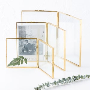 May include: A collection of clear glass and gold metal picture frames in various sizes. One frame displays a black and white photo with the word "PARIS" visible. A green fern and eucalyptus sprig are also present.