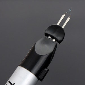 Könnte beinhalten: Ein schwarz-silberner Holzbrandstift mit Doppelspitzendesign. Der Stift hat einen schwarzen Griff und einen silbernen Schaft, auf dem das Wort "THR" teilweise zu sehen ist. Die Spitze hat zwei silberne Zinken mit einem kleinen blauen Draht.