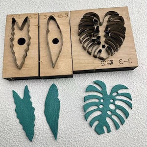 May include: A set of three wooden leaf-shaped leather cutting dies with metal blades, alongside three cut teal leather leaves. The dies are labeled with numbers and measurements. One die is a large monstera leaf.