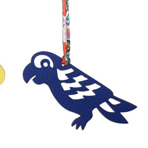 以下が含まれることがあります： 黄色と青色の2つのオウム型オーナメント。模様入りの布コードで吊り下げられています。オウムには切り抜きのディテールがあり、白い背景に配置されています。コードはねじれており、カラフルな花柄が特徴です。