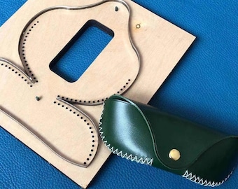 May include: A wooden craft template with a bird-like shape and a rectangular cutout, next to a dark green leather glasses case with white stitching and a gold button closure. The template has dotted lines for stitching.