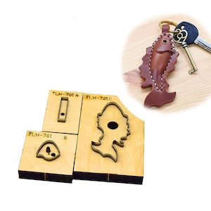 Può includere: Un portachiavi a forma di pesce in pelle marrone con cuciture bianche e un anello dorato. Accanto, tre fustelle in legno per lavori in pelle, etichettate TLH-701 A, B e C. Le fustelle servono per creare articoli in pelle a forma di pesce.