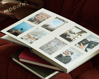 Puede incluir: Un álbum de fotos abierto con fotos de viajes descansa sobre dos libros cerrados en un sofá color óxido. El álbum muestra varias fotos de viajes, incluyendo escenas submarinas y vistas de ventanas de avión. Una manta marrón con texto blanco es parcialmente visible.