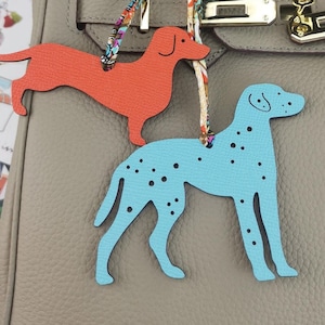 May include: Two leather dog-shaped bag charms. One is orange, resembling a dachshund, and the other is light blue with black spots, like a Dalmatian. Both charms are hanging from a bag with a gold clasp.