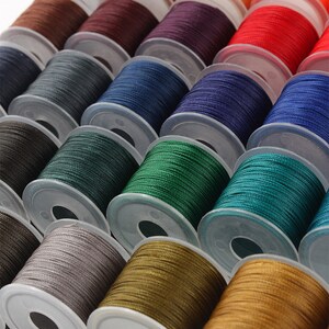 May include: Rows of colorful thread spools in shades of brown, orange, red, blue, green, and gold. The spools are neatly arranged, showcasing a variety of colors for sewing or crafting projects. The thread is tightly wound on clear plastic spools.