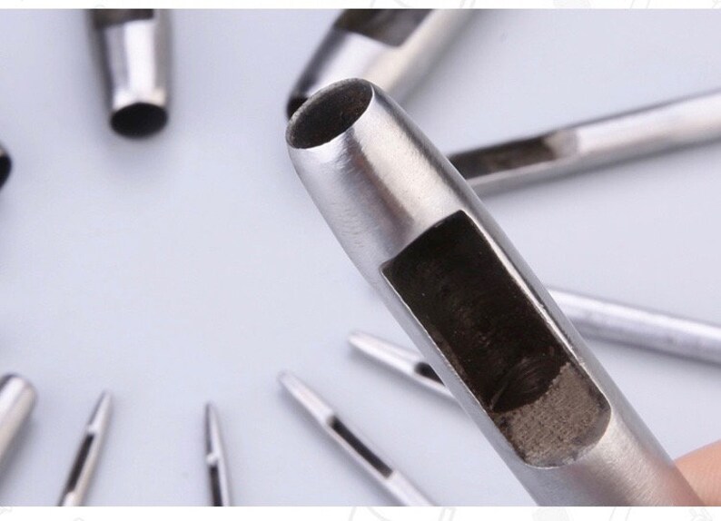 May include: Close-up of several silver-colored leather hole punches. The tools have a cylindrical shape with a hollow center and a rectangular opening on the side. The tools are designed for making holes in leather or similar materials.