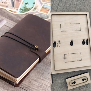 Op de afbeelding: Een bruin leren notitieboek met een zwarte elastische sluiting en een messing knoopsluiting. Het notitieboek ligt naast een houten stanssjabloon met verschillende vormen en uitsparingen, waaronder rechthoeken en druppels. Het notitieboek is ongeveer 15 cm hoog.