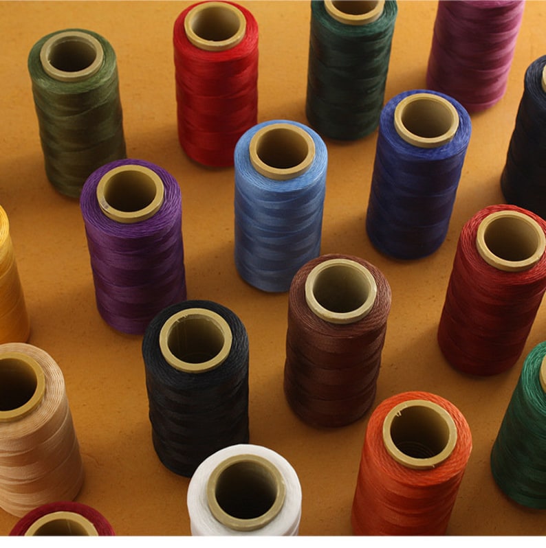 May include: A collection of spools of thread in various colors, including red, blue, green, and purple. The spools are cylindrical with a cardboard core, arranged on a brown surface. The threads appear to be tightly wound and ready for use.