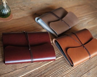 Könnte beinhalten: Drei Lederjournale in den Farbtönen Burgunderrot, Beige und Braun, jeweils mit einer Lederschnur befestigt. Die Journale sind auf einer Holzoberfläche angeordnet und zeigen ihre verschiedenen Farben und Texturen. Das Design ist schlicht und elegant.