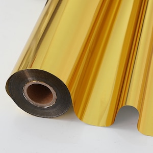 May include: A roll of gold foil, partially unrolled, showing a metallic surface. The foil is a bright, reflective gold colour, with a smooth, glossy finish. The roll is on a white background.