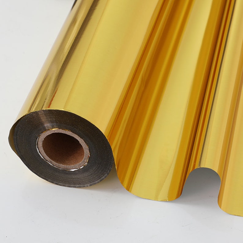 May include: A roll of gold foil, partially unrolled, showing a metallic surface. The foil is a bright, reflective gold colour, with a smooth, glossy finish. The roll is on a white background.