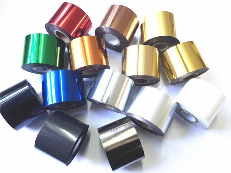 May include: An assortment of metallic foil rolls in colours such as red, gold, silver, green, and blue. The rolls are tightly wound and have a shiny, reflective surface. These foils are likely used for crafting or printing projects.