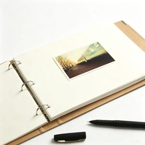 May include: An open ring-bound scrapbook with a tan cover and cream-colored pages. A rectangular photograph showing a train car on a platform is attached to the center of the right page. A black ballpoint pen and its cap lie nearby.
