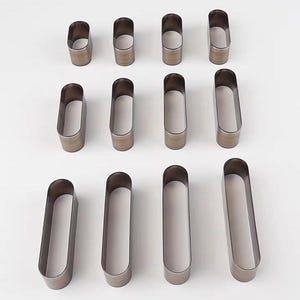 Könnte beinhalten: Eine Ansammlung von oval geformten Metall-Lederhandwerkzeugen, die auf einer weißen Oberfläche angeordnet sind. Die Werkzeuge sind in verschiedenen Größen erhältlich und haben eine abgerundete Oberseite und einen hohlen Innenraum. Das Metall hat ein dunkles, gealtertes Aussehen.