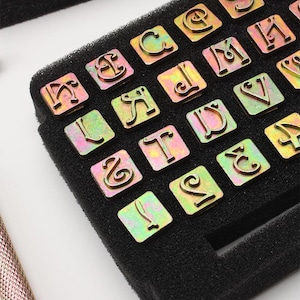 May include: A set of metal letter stamps arranged in a black foam insert. The stamps are in a variety of fonts and sizes, with a metallic finish. The letters are in a variety of fonts and sizes, with a metallic finish.