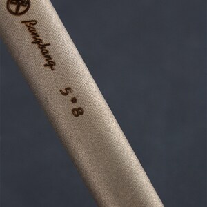 May include: Close-up of a beige cylindrical object with the text "Bangbang 5*8" in a dark brown colour. The object has a textured surface and a logo at the top. The background is a dark grey colour.