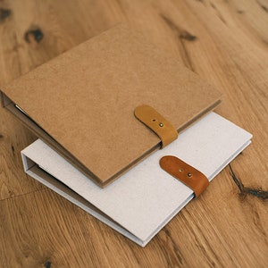 May include: Two square photo albums, one tan and one off-white, are stacked on a wooden surface. Each album has a leather strap closure with a metal rivet. The albums are likely for storing photographs or other keepsakes.