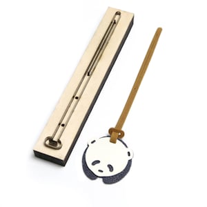 Puede incluir: Un troquel de madera con un canal largo y estrecho y un llavero de cuero con forma de panda. El llavero presenta una cara de panda blanca con detalles negros y un respaldo azul oscuro. El troquel es de color marrón claro.