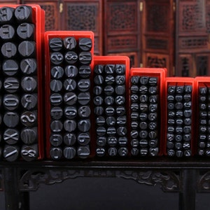 Puede incluir: Un conjunto de sellos negros de letras y n&uacute;meros en cajas de pl&aacute;stico rojo. Los sellos est&aacute;n dispuestos en filas y columnas, cada uno con un car&aacute;cter diferente. Las cajas son de diferentes tama&ntilde;os, lo que sugiere un juego completo de alfabeto y n&uacute;meros.