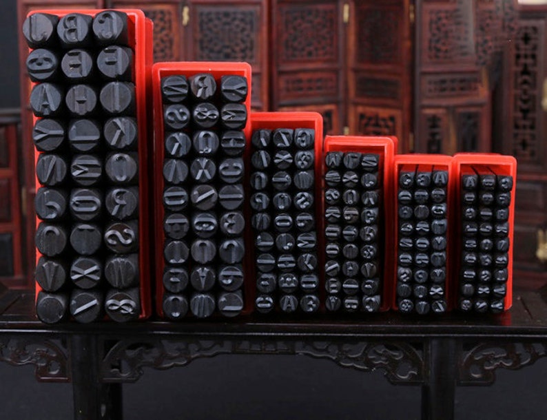 Puede incluir: Un conjunto de sellos negros de letras y n&uacute;meros en cajas de pl&aacute;stico rojo. Los sellos est&aacute;n dispuestos en filas y columnas, cada uno con un car&aacute;cter diferente. Las cajas son de diferentes tama&ntilde;os, lo que sugiere un juego completo de alfabeto y n&uacute;meros.