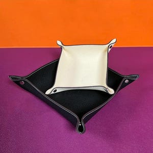 May include: Two leather catchall trays. The larger tray is black with white stitching and a cream-colored interior. The smaller tray is cream-colored with a dark brown trim. Both trays have a square shape with curved edges.