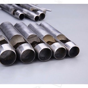 May include: A collection of silver-colored metal hole punches, arranged on a light gray surface. The punches are cylindrical with a hollow interior and a curved cutout on the side. Several other punches are in the background.