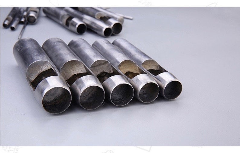 May include: A collection of silver-colored metal hole punches, arranged on a light gray surface. The punches are cylindrical with a hollow interior and a curved cutout on the side. Several other punches are in the background.