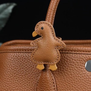 Puede incluir: Un bolso de mano de cuero marrón con un charm en forma de pato. El charm está hecho de cuero marrón con patas y pico amarillos, y está cosido alrededor de los bordes. El bolso tiene una superficie texturizada y un asa.