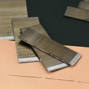May include: Close-up of leatherworking tools, including several rectangular metal blades with sharp edges, resting on a piece of tan leather with a stitched line. The blades have a wood-grain appearance.