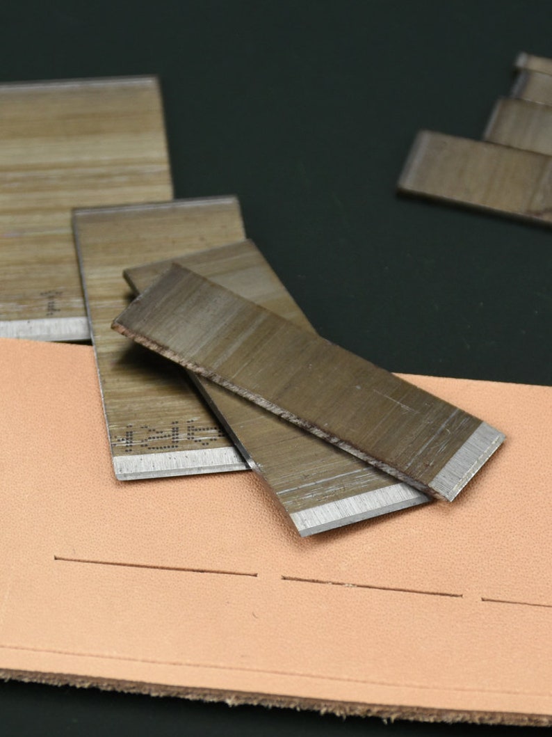 May include: Close-up of leatherworking tools, including several rectangular metal blades with sharp edges, resting on a piece of tan leather with a stitched line. The blades have a wood-grain appearance.