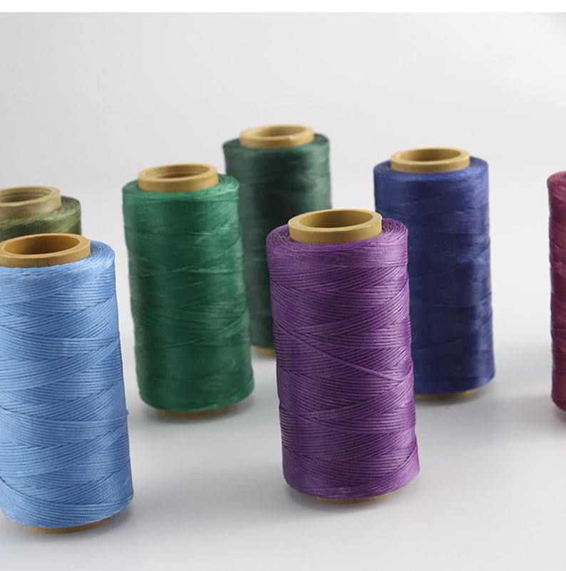 May include: A collection of colorful spools of thread in various shades, including blue, green, purple, and burgundy. Each spool is cylindrical, with the thread neatly wound around a cardboard core. The threads are tightly spun, suggesting durability.