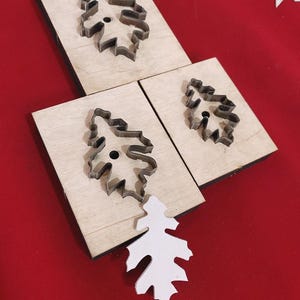 May include: Three wooden blocks with metal leaf-shaped cutouts, and a white leather leaf on a red surface. The cutouts are for crafting or leatherwork. The image shows the tools and a finished product.