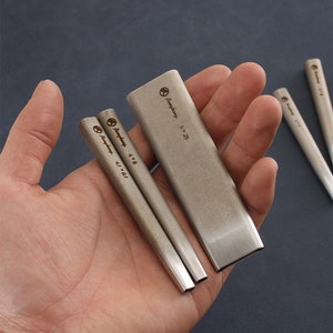 May include: A collection of silver woodworking chisels in different sizes. The chisels are rectangular with angled edges and size markings. The image displays the chisels against a dark backdrop.