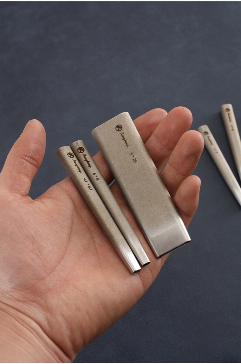 May include: A collection of silver woodworking chisels in different sizes. The chisels are rectangular with angled edges and size markings. The image displays the chisels against a dark backdrop.