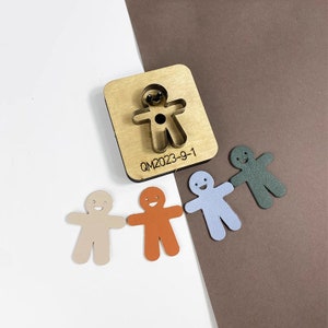 May include: A wooden die-cut tool with a metal gingerbread man shape, alongside four cut-out gingerbread man shapes in beige, orange, blue, and green. The tool has the text "QM2023-9-1" on it.
